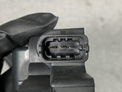 Close-up of a black three-pin automotive electrical connector with gold-plated pins held by a gloved hand (connector facing camera). Alternative: close-up of the mating end showing three gold pins.