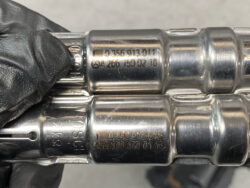 Close-up of two shiny metal automotive components with stamped part numbers, held by a gloved hand.