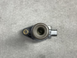 Top-down view of a cylindrical automotive sensor or connector with a circular opening, mounted on a black plastic housing on a gray surface.
