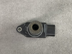 Close-up of a black automotive electrical component with a circular metal ring and a rectangular connector, placed on a gray surface.