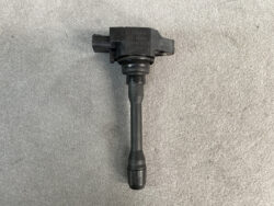 Black ignition coil with a rubber boot and electrical connector, laid on a gray surface (car engine part).