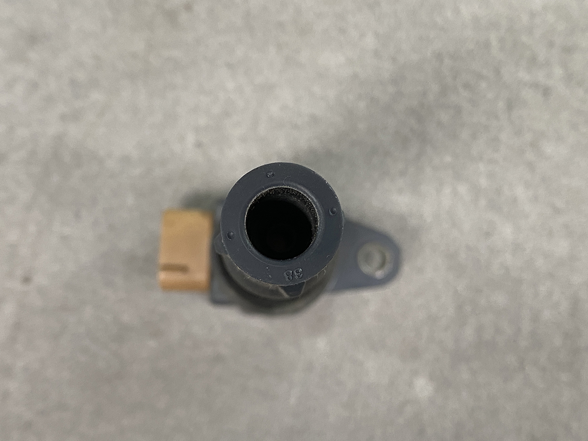 SUZ-COIL-DL3_5 SUZUKI Genuine IGNITION COIL / DL3 / 3731 / K12B, K6A, R06A Engine » NZ.95 » JDM-PARTS NZ » JDM-PARTS NZ Top-down view of a small gray plastic automotive sensor with a circular hollow opening and a side brown electrical connector, mounted on a metal surface. SUZUKI Genuine IGNITION COIL / DL3 / 3731 / K12B, K6A, R06A Engine » JDM-PARTS NZ