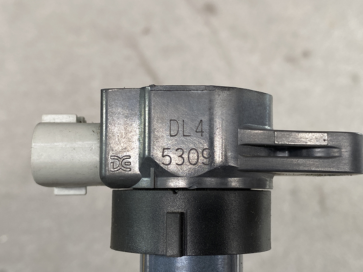 SUZ-COIL-DL4_2 SUZUKI Genuine IGNITION COIL / DL4 / 5309 / R06A Engine » NZ.95 » JDM-PARTS NZ » JDM-PARTS NZ Close-up of a gray metal mechanical component with the stamped code DL4 5309 and a dark circular clamp around a blue rod, on a light background. SUZUKI Genuine IGNITION COIL / DL4 / 5309 / R06A Engine » JDM-PARTS NZ