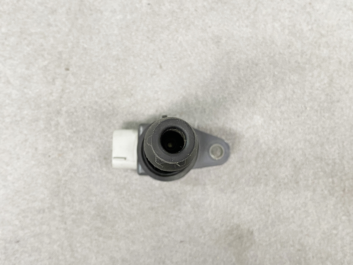 SUZ-COIL-DL4_5 SUZUKI Genuine IGNITION COIL / DL4 / 5309 / R06A Engine » NZ.95 » JDM-PARTS NZ » JDM-PARTS NZ Close-up of a small round camera lens in a black housing mounted on a gray bracket, viewed from above on a concrete surface. SUZUKI Genuine IGNITION COIL / DL4 / 5309 / R06A Engine » JDM-PARTS NZ