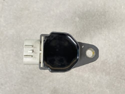 Automotive sensor module with a white electrical connector on the left and a mounting tab with a hole on the right, viewed from above on a gray surface.