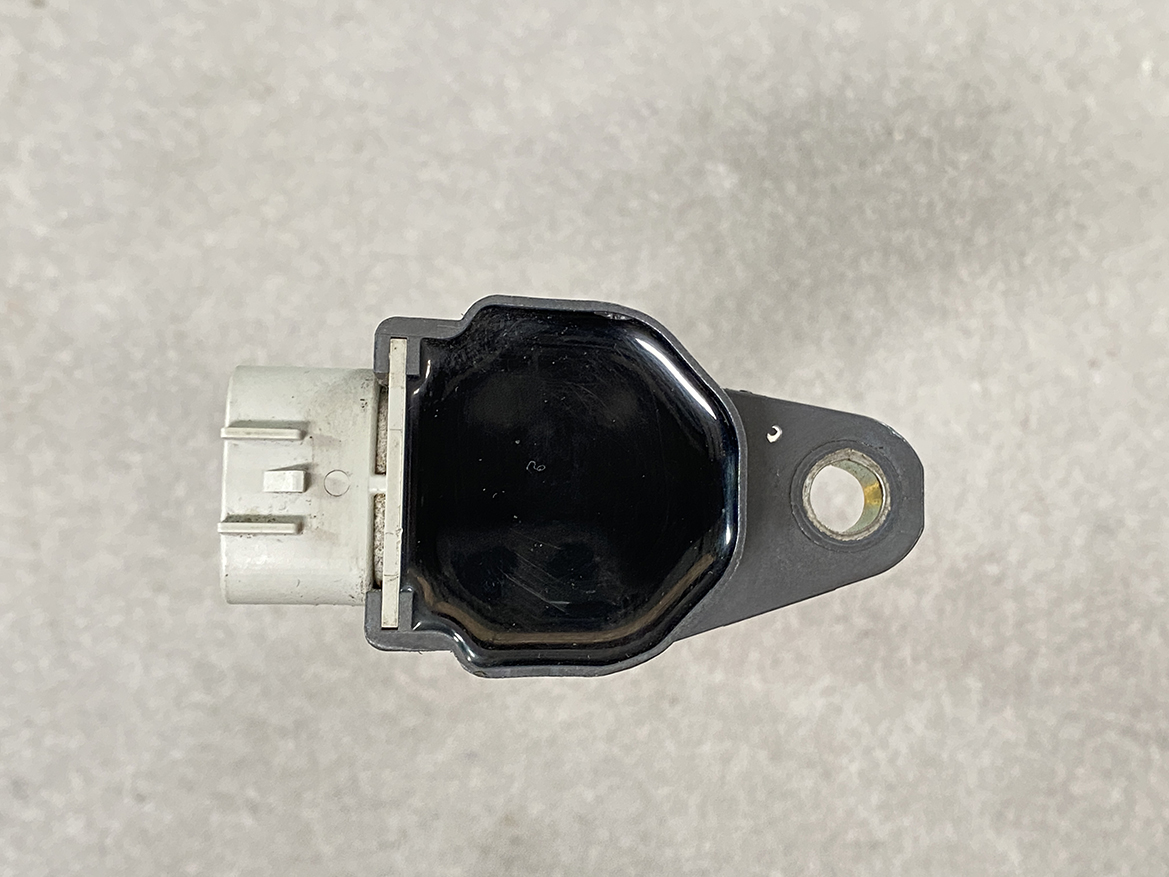 SUZ-COIL-DL4_6 SUZUKI Genuine IGNITION COIL / DL4 / 5309 / R06A Engine » NZ.95 » JDM-PARTS NZ » JDM-PARTS NZ Automotive sensor module with a white electrical connector on the left and a mounting tab with a hole on the right, viewed from above on a gray surface. SUZUKI Genuine IGNITION COIL / DL4 / 5309 / R06A Engine » JDM-PARTS NZ