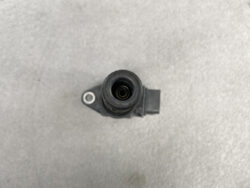 Close-up of a black automotive sensor housing with a circular inlet and a side mounting tab on a gray surface.