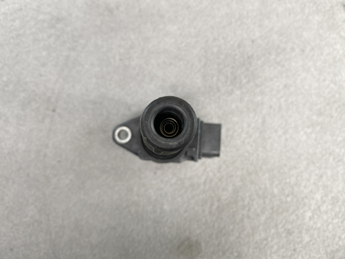 SUZ-COIL-FK0508_5 SUZUKI Genuine IGNITION COIL FK0508 / R06A, K12B Engine » NZ.95 » JDM-PARTS NZ » JDM-PARTS NZ Close-up of a black automotive sensor housing with a circular inlet and a side mounting tab on a gray surface. SUZUKI Genuine IGNITION COIL FK0508 / R06A, K12B Engine » JDM-PARTS NZ