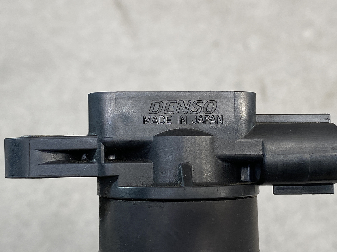 SUZ-COIL-NL2_3 SUZUKI Genuine IGNITION COIL DENSO NL2 099700 / R06A, K6A Engine » NZ.95 » JDM-PARTS NZ » JDM-PARTS NZ Close-up of a black plastic DENSO automotive component with the engraving 'DENSO MADE IN JAPAN' visible, mounted on a cylindrical surface. SUZUKI Genuine IGNITION COIL DENSO NL2 099700 / R06A, K6A Engine » JDM-PARTS NZ