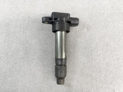 Automotive ignition coil with black plastic top and metal cylindrical body on a gray textured surface.