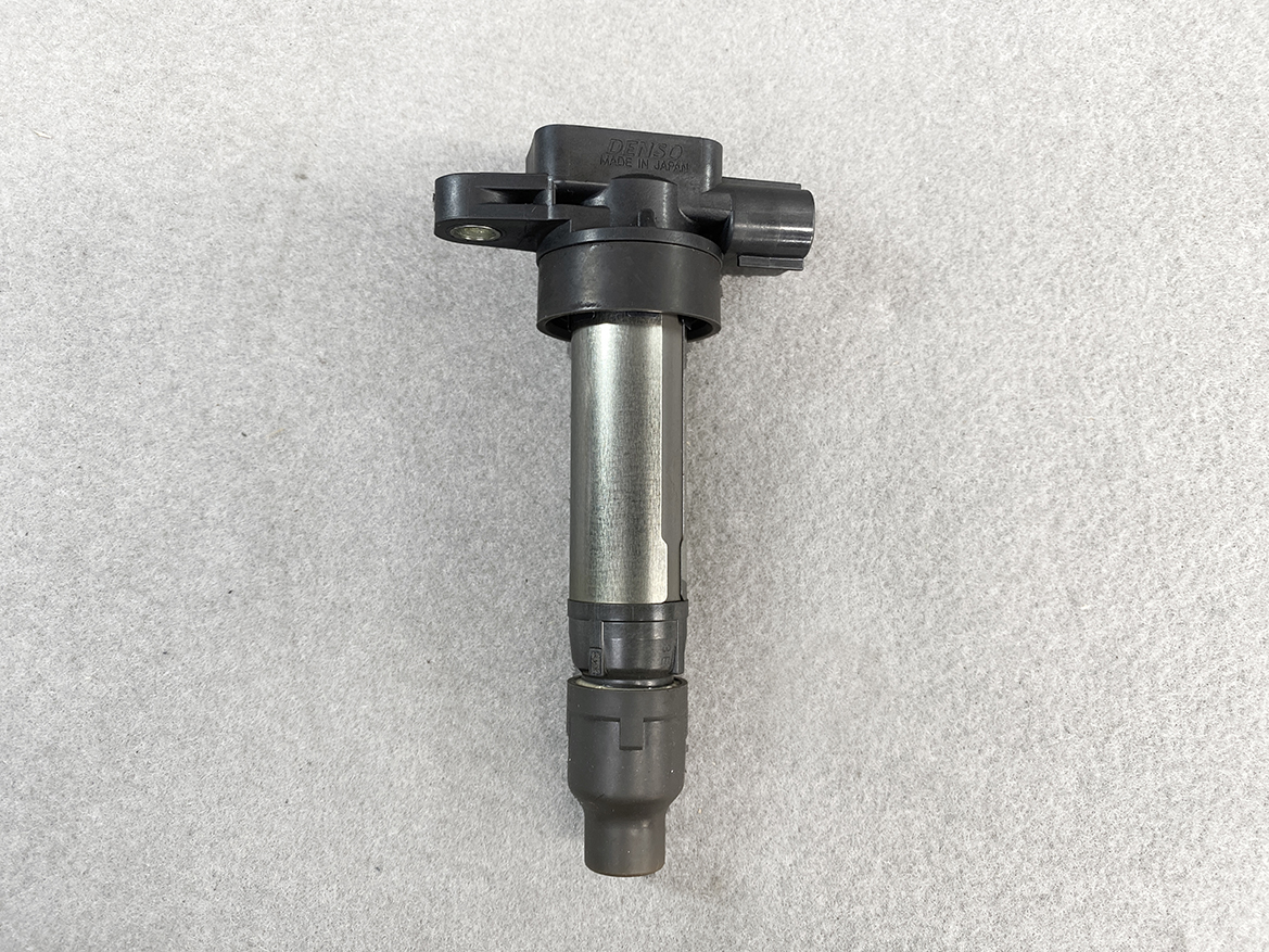 SUZ-COIL-NL2_5 SUZUKI Genuine IGNITION COIL DENSO NL2 099700 / R06A, K6A Engine » NZ.95 » JDM-PARTS NZ » JDM-PARTS NZ Automotive ignition coil with black plastic top and metal cylindrical body on a gray textured surface. SUZUKI Genuine IGNITION COIL DENSO NL2 099700 / R06A, K6A Engine » JDM-PARTS NZ