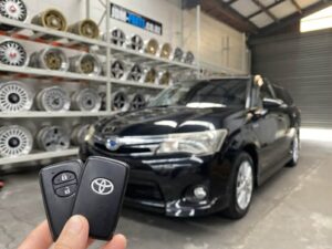 Person holding Toyota car key fob in front of a black sedan inside a shop with wheels on racks in the background.