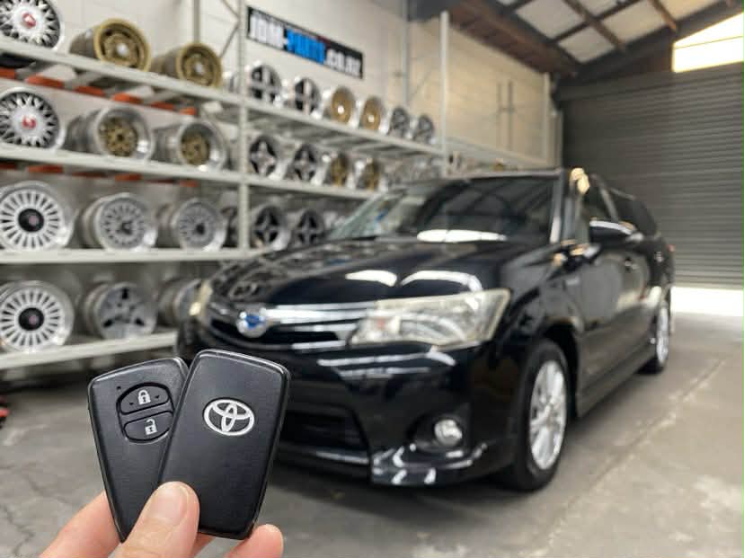 Person holding Toyota car key fob in front of a black sedan inside a shop with wheels on racks in the background. TOYOTA AXIO Replacement Key » JDM-PARTS NZ