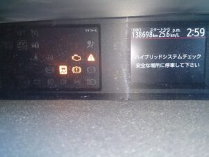 Car dashboard with illuminated warning icons; odometer 138,698 km, efficiency 25.6 km/L, time 2:59. Japanese safety message on right screen.
