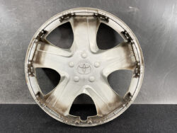 Circular gray Toyota hubcap with a five-spoke design, showing the Toyota emblem in the center, resting on a dark surface and looking dusty.