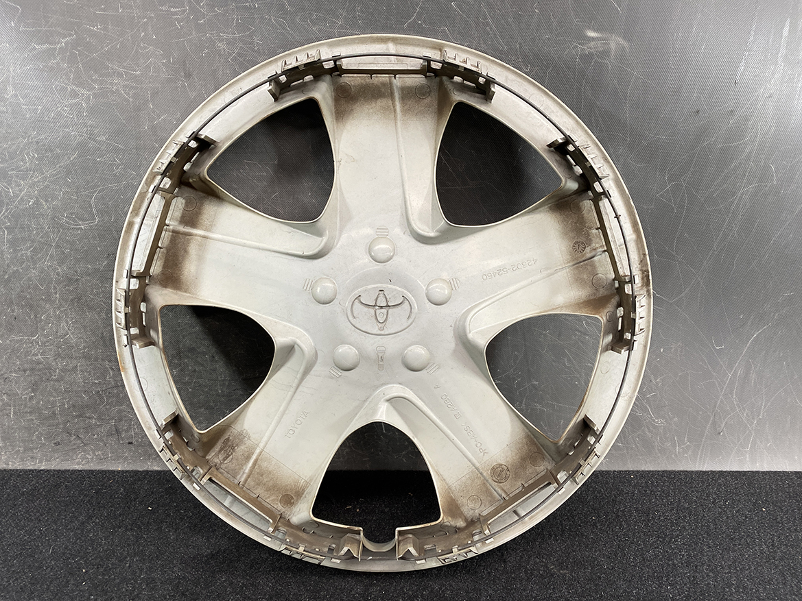 B392_3 TOYOTA RACTIS NSP120, NCP120 Genuine 16inch WHEEL CAP / HUB CAP 42602-52460 » NZ.95 » JDM-PARTS NZ » JDM-PARTS NZ Circular gray Toyota hubcap with a five-spoke design, showing the Toyota emblem in the center, resting on a dark surface and looking dusty. TOYOTA RACTIS NSP120, NCP120 Genuine 16inch WHEEL CAP / HUB CAP 42602-52460 » JDM-PARTS NZ