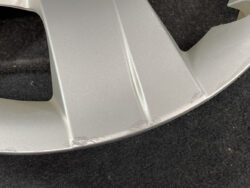 Silver alloy wheel with noticeable scrape marks along the lower lip rim area against a dark background, indicating minor damage.