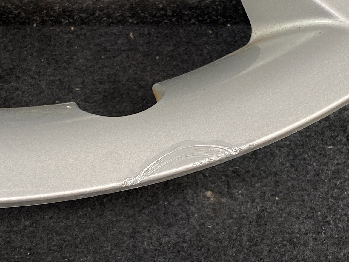 B393_5 TOYOTA RACTIS NSP120, NCP120 Genuine 16inch WHEEL CAP / HUB CAP 42602-52460 » NZ.95 » JDM-PARTS NZ » JDM-PARTS NZ Close-up of a silver curved metal surface with a scrape and scuff marks near a notch, on a dark background. TOYOTA RACTIS NSP120, NCP120 Genuine 16inch WHEEL CAP / HUB CAP 42602-52460 » JDM-PARTS NZ