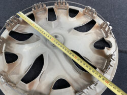 Circular plastic wheel or hubcap with multiple triangular cutouts, with a yellow measuring tape laid diagonally across it.