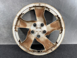 Rusty metal car wheel rim with five spokes and central lug holes, resting on a dark surface.