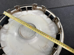 Round gray plastic car hubcap on a dark surface with a tape measure stretched diagonally across it, showing the size measurement.
