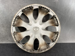 Round silver Toyota hubcap with ten spokes, dusty and scuffed, against a gray wall.