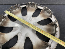 Round metal car wheel cover with eight spoke-like openings and a yellow tape measure laid across it