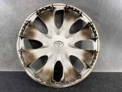 Used silver Toyota hubcap (wheel cover) with logo, dusty and worn, resting on a dark floor against a textured wall.