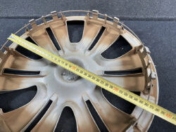 Tan plastic car wheel hubcap laid flat on a dark surface with a yellow measuring tape stretched diagonally across it, showing the diameter measurement approach.