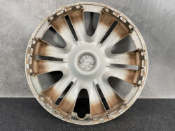 Round alloy wheel hubcap with multiple spokes and a central cap, dusty with rust-colored stains, resting on a dark surface