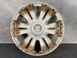 Silver car wheel rim with multiple spokes, showing rust and wear, set against a dark textured background