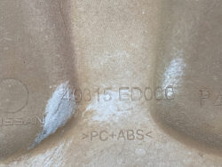 Close-up of a brown plastic automotive part with stamped markings reading 40315 ED000 and PC+ABS; the word NISSAN is partially visible in the corner.