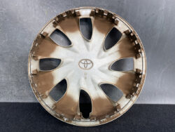 Dirty Toyota hubcap wheel cover with multiple spokes, placed on a dark surface; round, beige-brown plastic with the Toyota emblem in the center