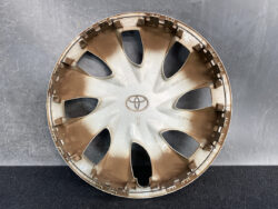 Round beige Toyota hubcap with brown dirt, central Toyota logo, and multiple clip tabs around the rim against a dark background.