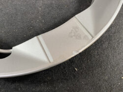 Close-up of a light gray curved plastic piece with two subtle ridges and small scuff marks on a dark carpeted surface.