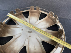 Brown plastic wheel hub cap with a yellow measuring tape laid across it, showing about 300 mm (30 cm) length, placed on a dark surface.