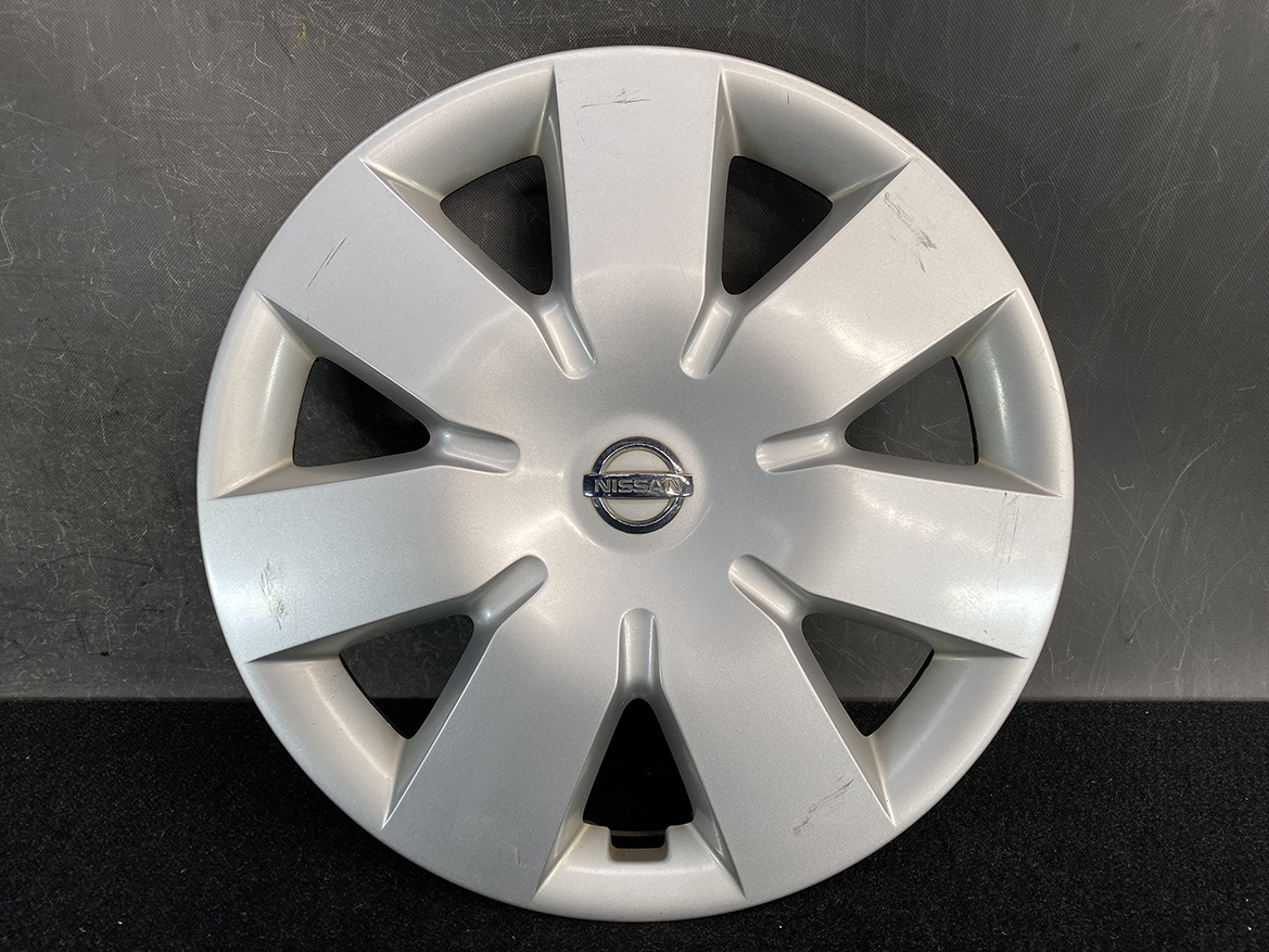 B438_2 NISSAN NOTE E11 Genuine 14inch WHEEL CAP / HUB CAP x1 40315 1U600 x1 » NZ.95 » JDM-PARTS NZ » JDM-PARTS NZ Silver Nissan hubcap with eight spokes and a central Nissan emblem, showing light surface scratches on the rim side trims while resting on a dark backdrop. NISSAN NOTE E11 Genuine 14inch WHEEL CAP / HUB CAP x1 40315 1U600 x1 » JDM-PARTS NZ