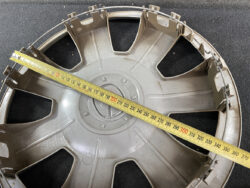 Plastic car wheel cover with radial spokes, laid on a dark surface, a tape measure stretched across it to show size.