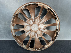 Brown plastic car wheel hubcap with twelve spokes laid face up, showing scuffs and wear on a dark surface.