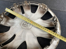 Diagonal measuring tape laid across a silver car wheel rim on a dark surface, showing measurement marks up to ~300 mm (about 12 inches). This image highlights sizing of the hubcap.