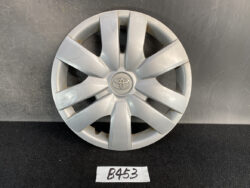 Silver Toyota hubcap with an eight-spoke star design, showing light scratches, resting on a dark surface with a small handwritten tag labeled B453