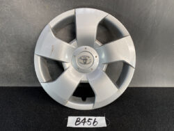 Front view of a silver Toyota wheel cover with visible scratches and a damaged bottom edge, placed on a dark surface with a labeled tag reading B456.