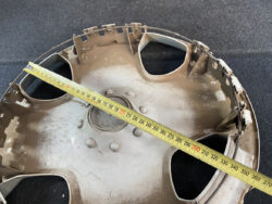 Close-up of a circular metal car wheel hubcap with broken edges, a measuring tape stretched diagonally across for scale on a dark surface.
