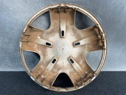 Rusty brown circular car hubcap with six spokes and a central hub, faded Honda logo, lying on a dark surface.
