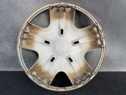 Dusty beige Honda hubcap with a six-spoke star pattern, photographed face-on against a dark textured ground surface.