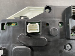 White multi-pin electrical connector on a black plastic panel, with gold contacts visible inside the housing