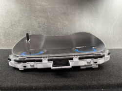 Unassembled car instrument cluster module with clear curved lens and two blue-accented gauge dials inside a white plastic housing behind a transparent cover