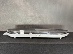 Long automotive interior trim panel with a curved black top, silver mounting clips, and a white base on a dark surface.