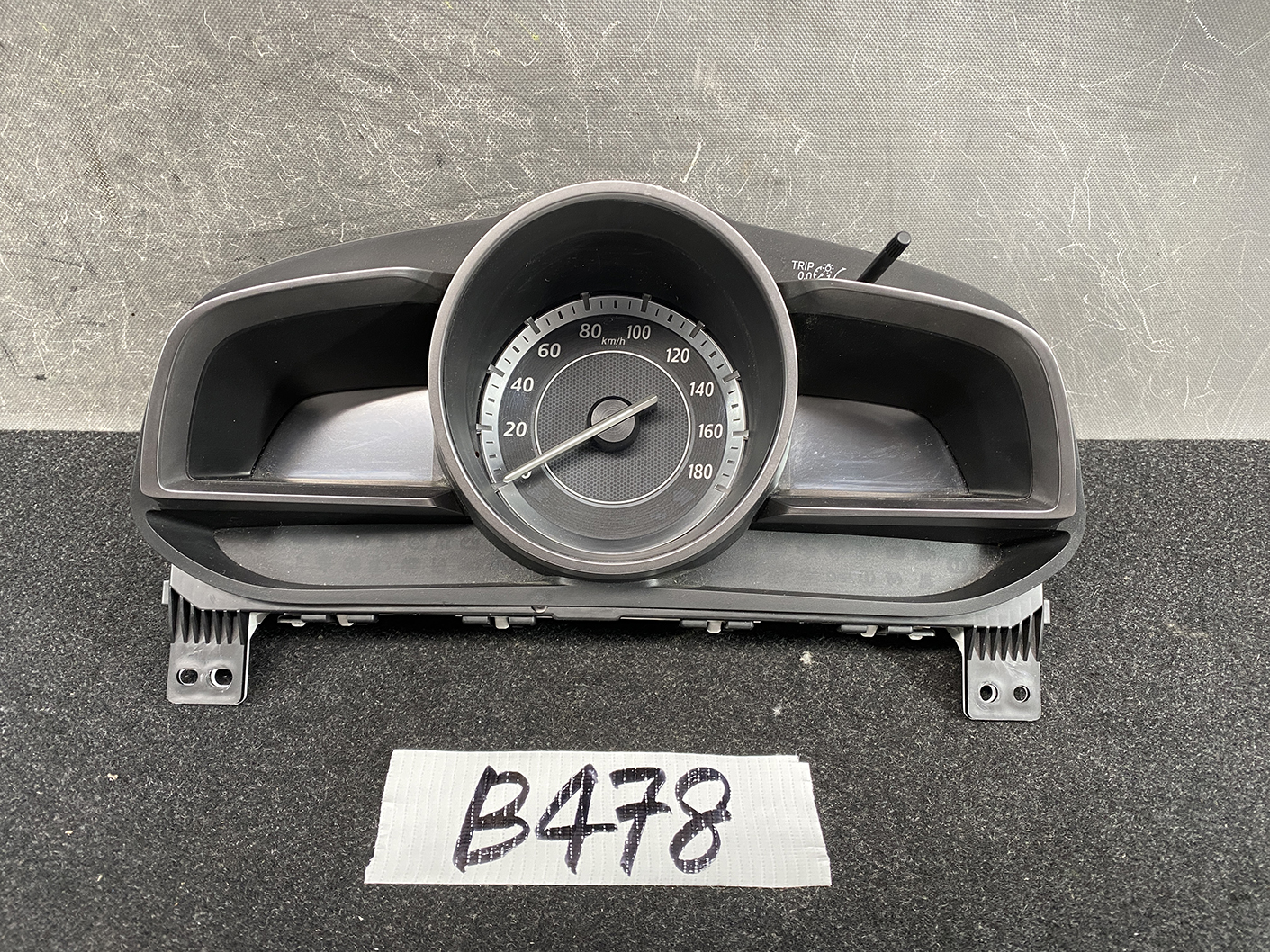 B478_2 MAZDA DEMIO CX-3 Genuine Instrument Cluster K1054 / D09M 55 430 » NZ1.95 » JDM-PARTS NZ » JDM-PARTS NZ Circular car speedometer displaying 0–180 km/h inside a black plastic housing on a grey surface. MAZDA DEMIO CX-3 Genuine Instrument Cluster K1054 / D09M 55 430 » JDM-PARTS NZ