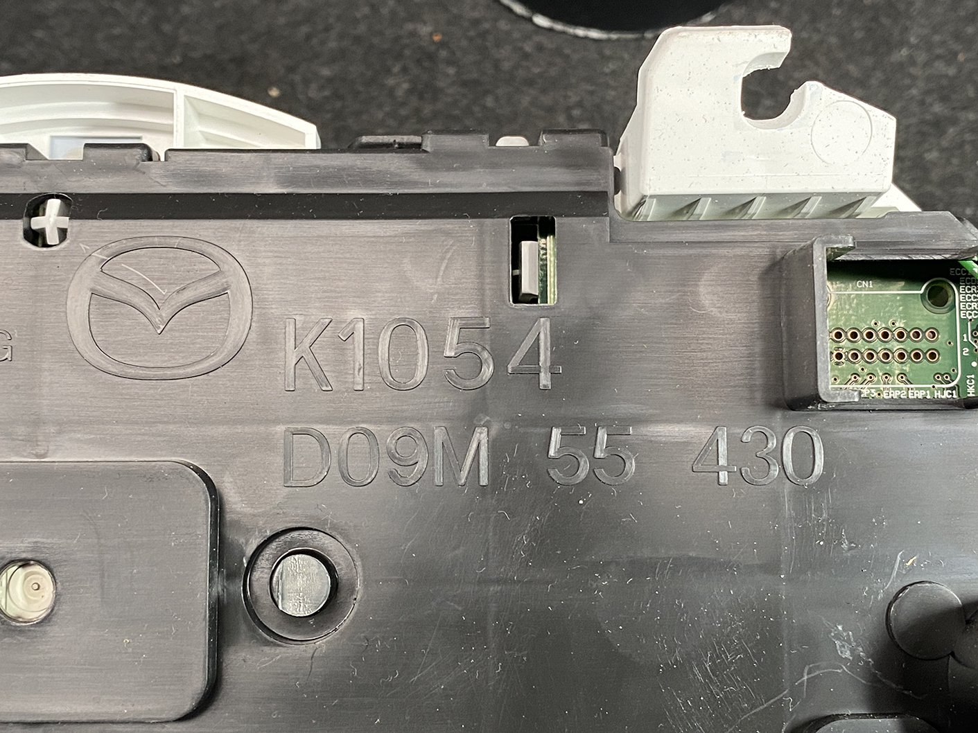 B478_4 MAZDA DEMIO CX-3 Genuine Instrument Cluster K1054 / D09M 55 430 » NZ1.95 » JDM-PARTS NZ » JDM-PARTS NZ Close-up of a car electronic control unit housing with a Mazda logo and part numbers K1054 D09M 55 430, exposing a small circuit board edge. MAZDA DEMIO CX-3 Genuine Instrument Cluster K1054 / D09M 55 430 » JDM-PARTS NZ